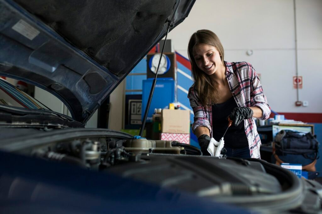 Young casual woman in shirt and gloves working with car in repair service checking engine oil level